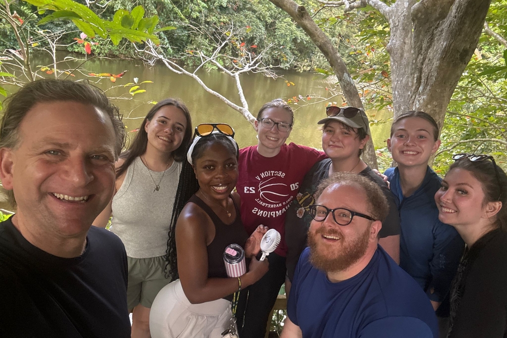 A group of students and two adults pose for a selfie beside a calm river surrounded by dense tropical trees and branches. Everyone is smiling, with some wearing glasses or sunglasses, and the group stands closely together on a shaded wooden overlook.