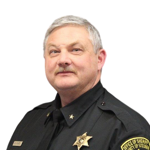 Official portrait of a uniformed sheriff's office official wearing a black dress uniform with badge and insignia, photographed against a light gray background.
